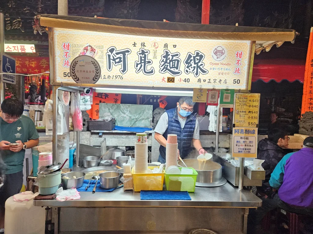士林廟口老店阿亮麵線是讓很多台北人從小吃到大的士林老字號美食，飄香夜市超過半世紀以上，至今依舊維持那一碗樸實、勾芡濃郁的大腸蚵仔麵線風味，堪稱士林夜市必吃小吃之一。無論是本地人還是旅客，只要走過士林廟口這家麵線攤位前，都會不自覺停下腳步聞香而來。