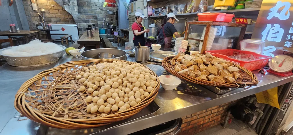 魚丸伯仔 這家傳承超過六十年的老店，必點組合就是魚丸湯配上豆干包。魚丸選用新鮮鯊魚漿製作，口感扎實彈牙且帶有淡淡鮮甜，咬下時能感受到魚漿的純粹；豆干包則是將油豆腐塞入特製肉燥後以魚漿封口，滷汁滲透進豆腐孔洞中，搭配獨門辣醬更是層次分明，是極具代表性的古早味。