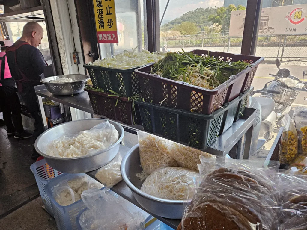 金榜麵館備料區展現巨量食材:金屬大盆裝滿雪白客家粄條,一旁籃子則堆積如山、份量驚人的豆芽菜與綠韭菜,印證其便宜大碗的美名。